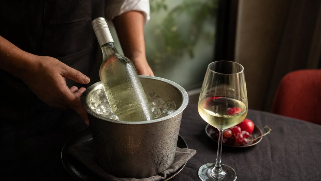 A single chilled white wine bottle resting in a metal ice bucket on a dark linen-covered table, beads of condensation catching soft window light. Beside it, one clear stemmed glass shows pale straw reflections with a faint red-toned garnish from nearby fruit, creating a calm, refreshing mood.