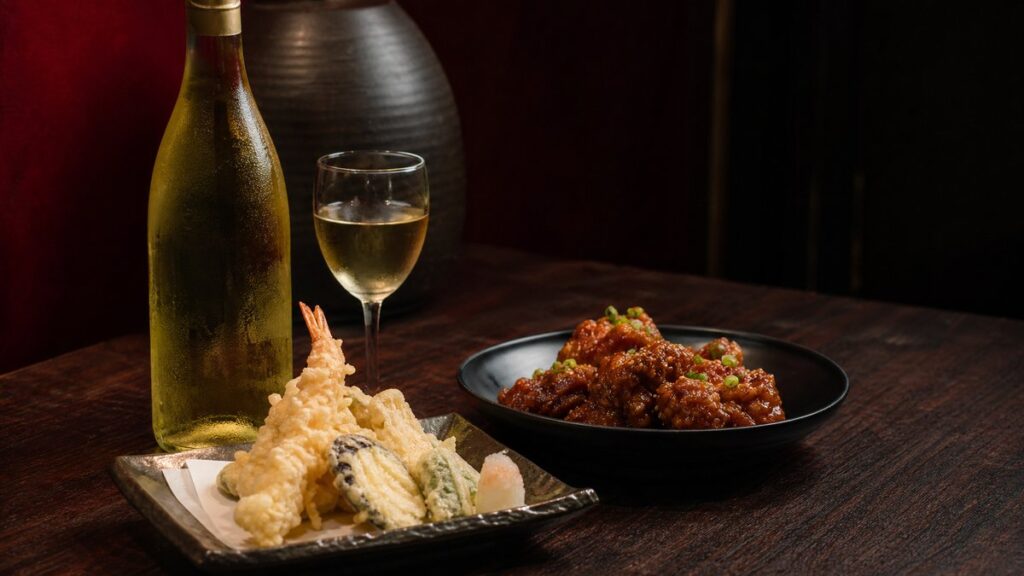 A professional editorial still life on a dark wooden counter: a chilled white wine bottle beside two different fried dishes, one plate of pale crisp tempura and another of glossy sweet-salty fried chicken. Soft side lighting catches the oil sheen and delicate bubbles in batter, with deep burgundy and muted black tones adding warmth and contrast.