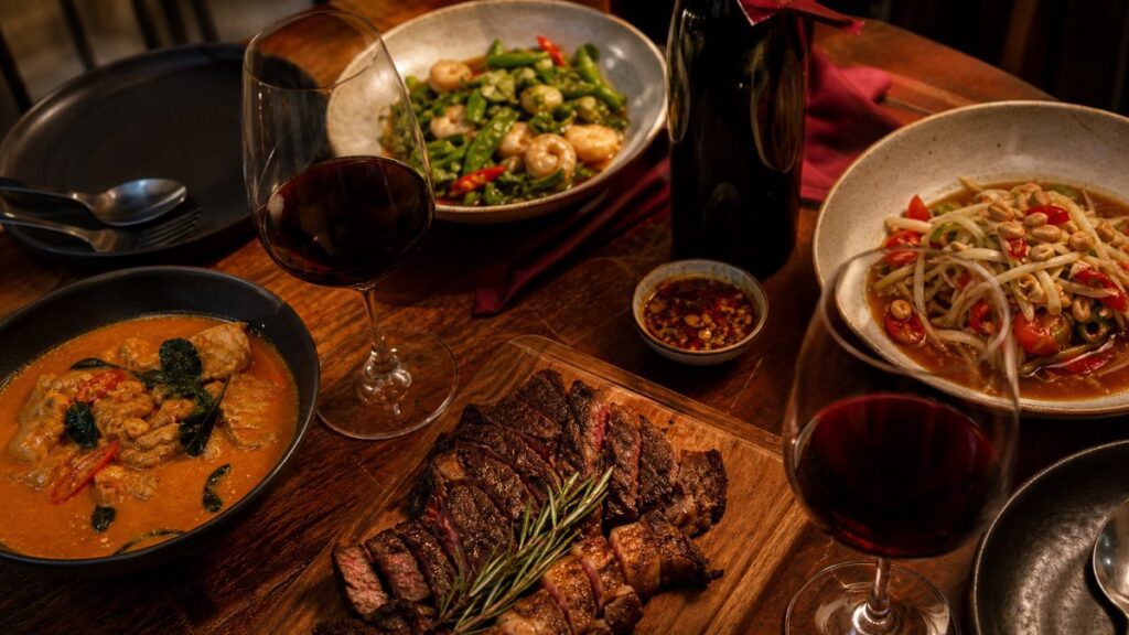 An overhead scene shows a simple dinner setting with grilled meat, spicy Thai dishes, and a bottle of red wine opened nearby, arranged naturally on a dining table. Warm evening light falls across the food, creating a lively, practical mood that suggests pairing wine to the meal, not the label.