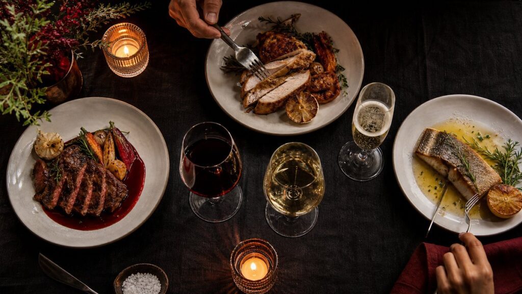 A refined still life on a dark linen table: three partially filled glasses of red wine, white wine, and sparkling wine arranged beside seared steak, roasted chicken, and buttered fish on plain plates. Warm candlelight and a deep burgundy accent in the sauce create a professional, intimate dinner mood.