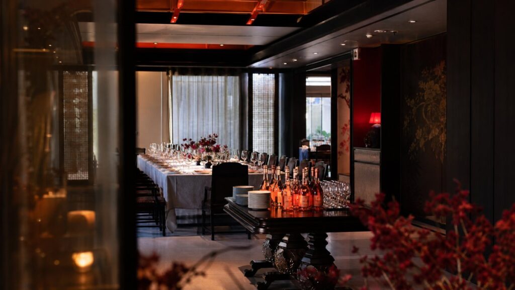 A wide restaurant private room prepared for a small reception, with a central table of rosé bottles, white plates, and neatly arranged chairs under gentle ambient lighting. The scene feels versatile and polished, with deep #0C0707 shadows, subtle ruby highlights, and an easygoing yet professional event mood.