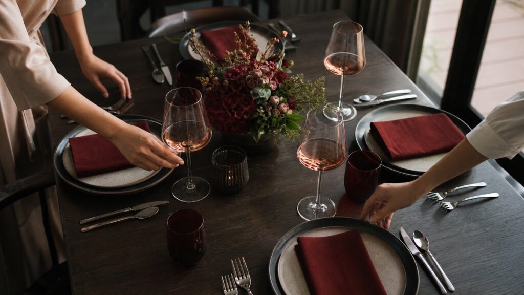 An overhead view of a formally set dinner table being finished with careful placement of rosé glasses, polished cutlery, and folded napkins by unseen hands. Soft evening light from a nearby window and dark tabletop shadows add a composed, upscale atmosphere, with muted red accents woven naturally into the scene.