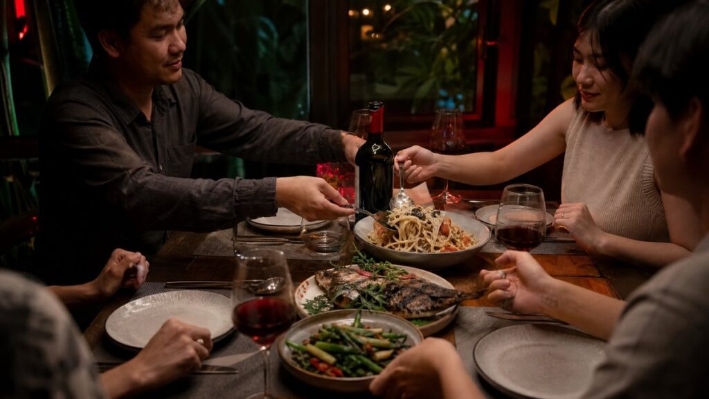 A wide restaurant environment with a modest dinner table for four by a softly lit window, where a single bottle of wine and shared plates of pasta, grilled fish, and vegetables anchor the scene. The atmosphere feels relaxed and welcoming, with subtle red reflections and dark shadows adding depth.