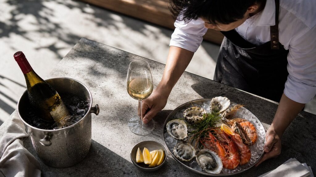 An overhead process scene showing a server carefully placing a stemmed white wine glass, a small bowl of lemon wedges, and a seafood platter on a lightly textured table. The wine sits in a bucket with condensation, and cool daylight emphasizes precision, freshness, and the quiet ritual of serving at the right moment.