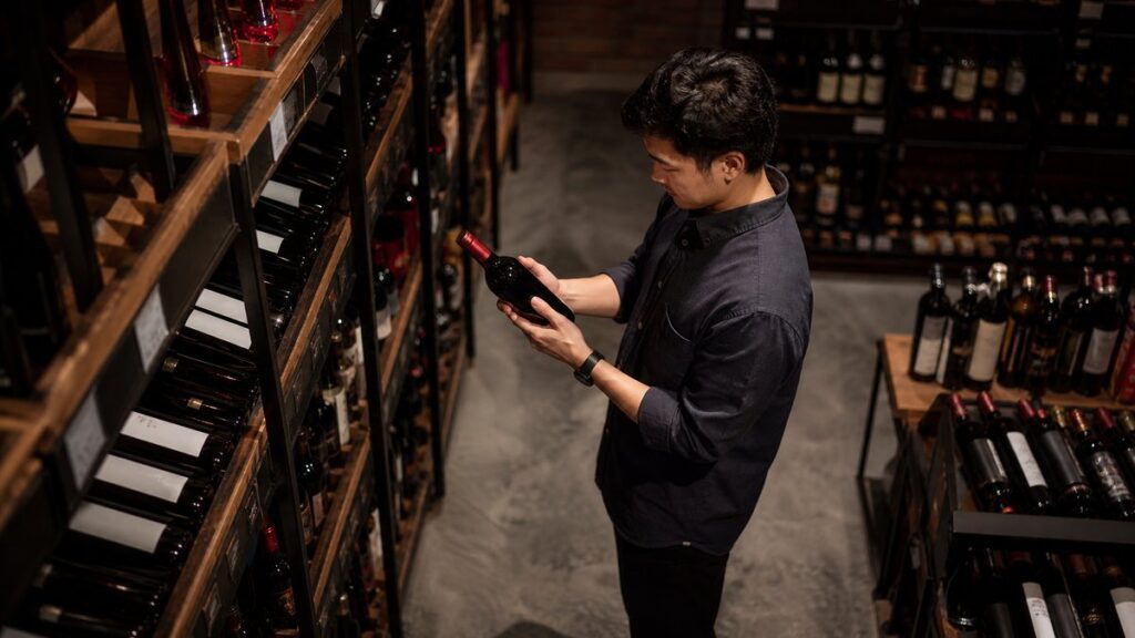 A wide view inside a modest wine shop shows a focused shopper standing among neatly stocked shelves, studying a single bottle in hand while the rest of the aisle fades softly behind. The lighting is calm and professional, with dark charcoal tones and faint red accents conveying thoughtful, trustworthy decision-making.