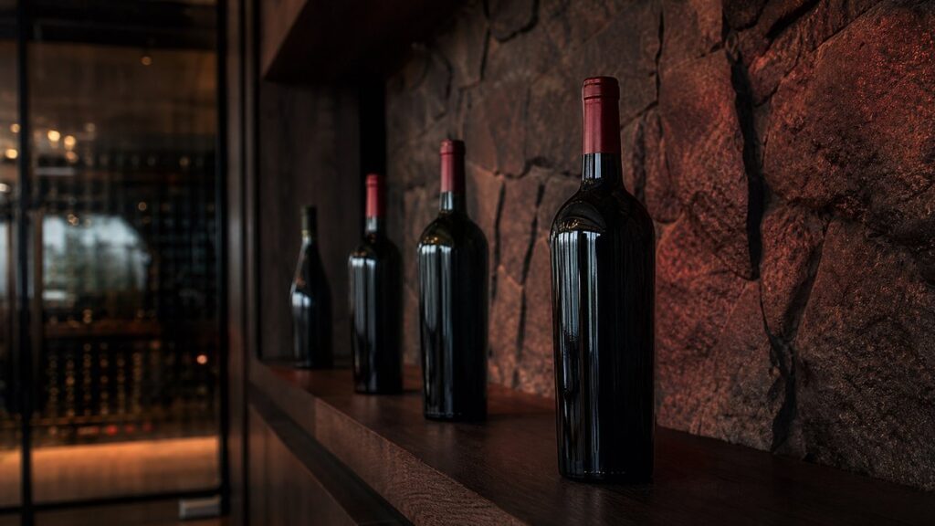 A wide cellar shelf with a few wine bottles spaced apart, one bottle pulled slightly forward as the primary subject. Soft ambient light washes the stone wall in deep burgundy and dark charcoal tones, creating a quiet, confident mood. The composition leaves generous negative space and feels professional.