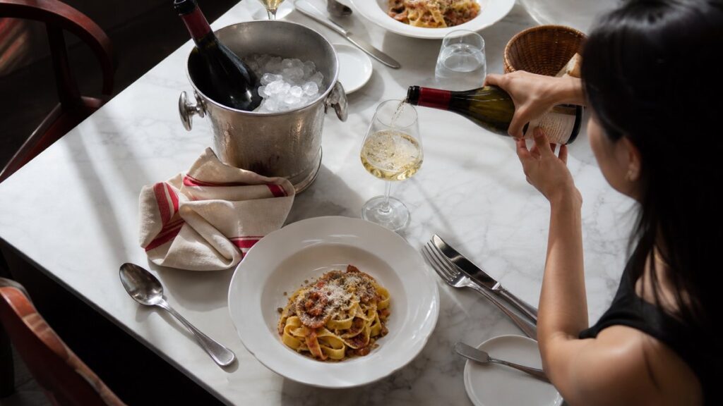 Overhead view of a clean dining setup: a bottle resting in an ice bucket, a precise pour into a tulip-shaped glass, and a plated pasta dish arranged beside it. Cool daylight from the side makes the wine look crisp and bright, with subtle red accents in a folded linen and warm reflections on polished tableware.