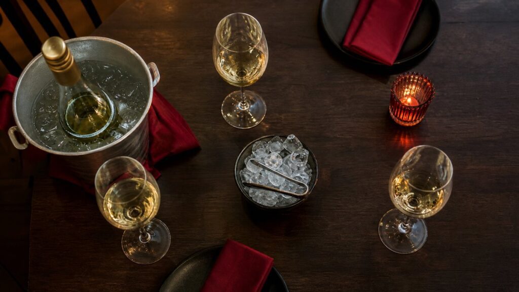 Overhead view of a simple dining table arranged with three wine glasses, a chilled white wine bucket, and a small bowl of ice, all placed with careful spacing on a dark wood surface. Subtle crimson highlights in napkins and candlelight add richness, while the composition feels restrained, polished, and professional.