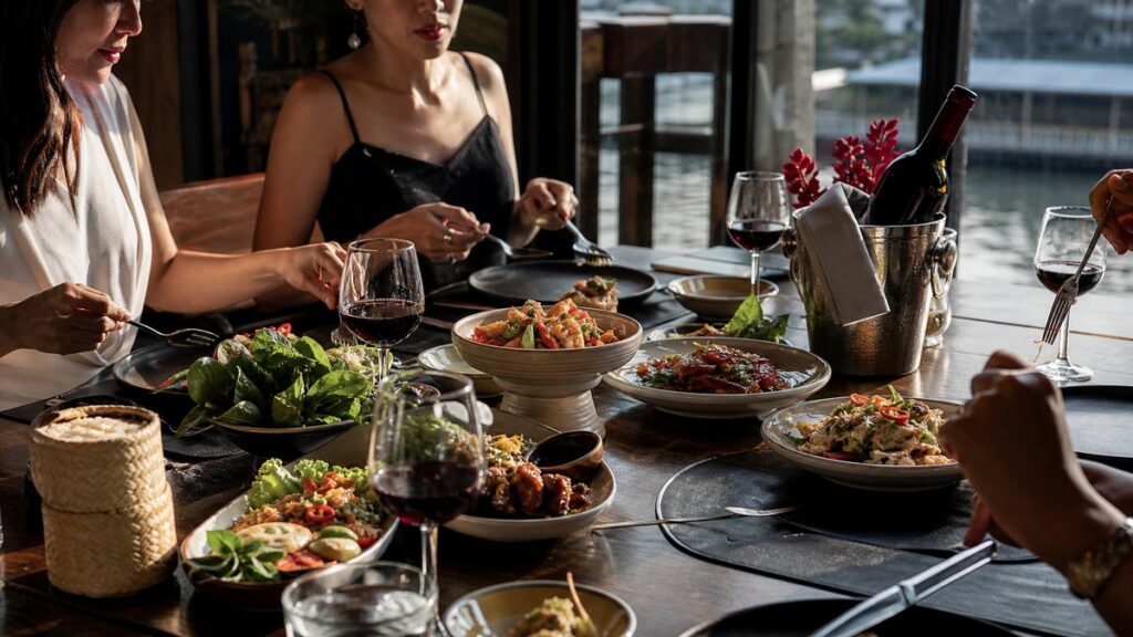A wide restaurant table set for an early evening meal, with Thai dishes centered around a chilled bottle of red wine in a simple bucket. Natural side lighting from a window highlights the food textures, while subtle red accents in garnishes and reflections add a polished, professional atmosphere.