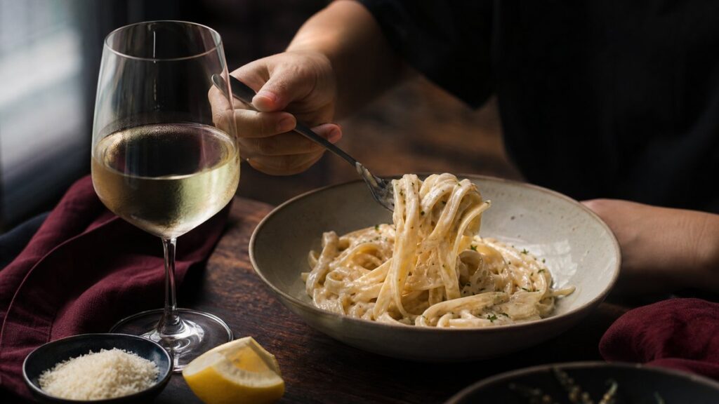 A tabletop still life of a bowl of creamy fettuccine beside a chilled glass of white wine, with a lemon wedge and a small dish of grated cheese nearby. Soft window light highlights the pasta’s sheen and the wine’s pale straw color, while deep burgundy napkins and dark shadows add a restrained, professional mood.