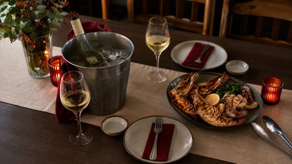 An overhead view of a neatly arranged dinner table with a wine bucket, chilled white wine, a sparkling bottle, small plates, tongs, and grilled seafood on a linen runner. Soft evening light falls evenly across the setup, and subtle red accents in napkins and candle glow create an elegant, practical atmosphere.