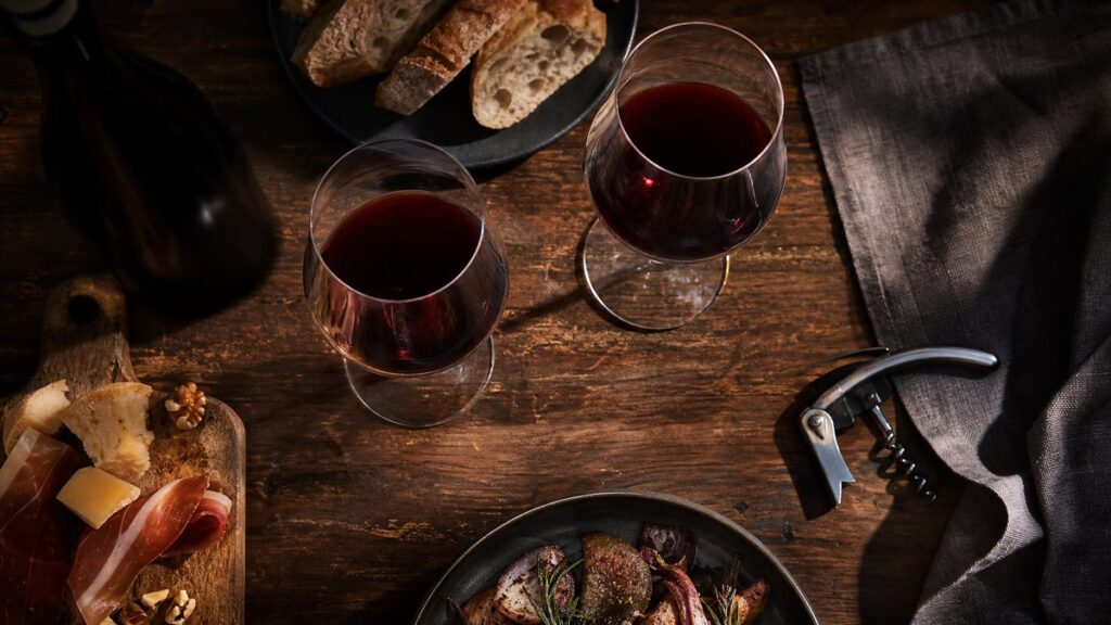 Overhead on a rustic wooden table, two unlabeled wine glasses sit beside simple food pairings, with an opened corkscrew and a modest meal in the frame. One bottle is implied only by its silhouette, keeping the scene grounded and realistic. Warm side lighting and restrained red highlights suggest price is not the deciding factor, creating a thoughtful, unpretentious atmosphere.