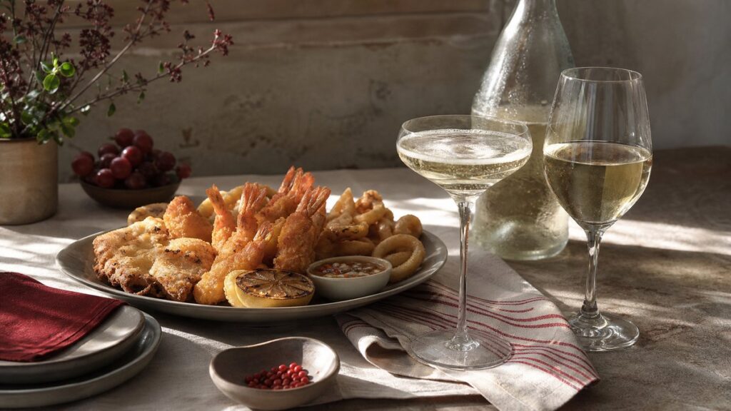 A wide kitchen-table environment showing a simple shared meal: a bottle of sparkling wine, a clear white wine, and a plate of assorted fried foods arranged naturally on linen and stoneware. Morning light falls across the scene, emphasizing crisp textures, light foam, and a calm everyday mood with restrained dark crimson accents in fruit and napkin details.