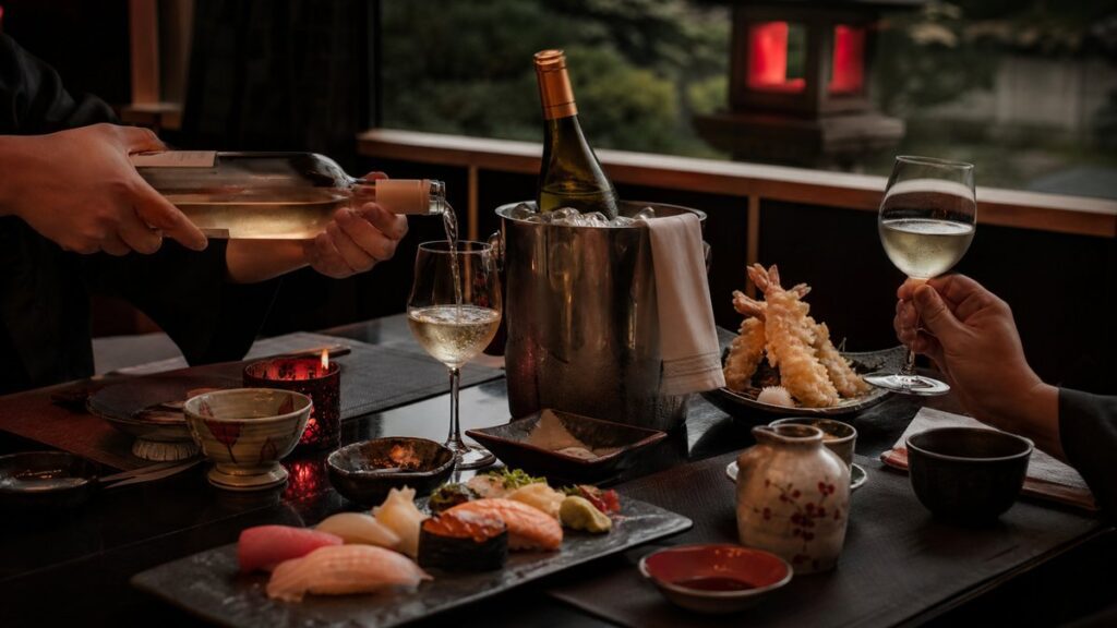 A wide dining-room scene shows a simple Japanese meal set for two on a low table near a window, with white wine chilled in an ice bucket and sushi, tempura, and ceramic dishes spaced neatly around it. Evening light washes the room in deep black and subtle red tones, giving an understated celebratory feeling.