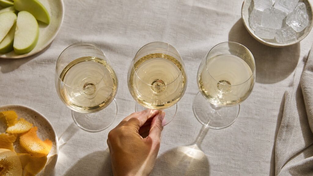 Overhead view of three different white wine glasses arranged on a linen-covered table, each showing a slightly different hue and clarity beside citrus peel, green apple slices, and a small bowl of ice. Natural daylight reveals contrast in texture and brightness, creating an analytical yet inviting editorial feel.