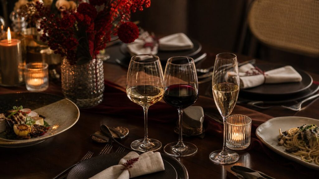 An overhead scene of a neatly arranged dinner setting for a special occasion, showing three different wine glasses near a table set with elegant but simple dishes, candles, and folded napkins. Warm evening light and muted red accents create a celebratory yet relaxed atmosphere, suggesting thoughtful choices for family, couple, or festive meals.