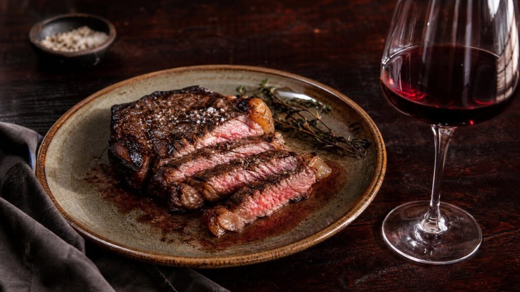 A close tabletop still life shows a sliced ribeye steak on a warm ceramic plate beside a half-filled red wine glass, with glossy meat juices and a faint charred crust catching soft side light. Deep burgundy reflections mingle with dark wood and linen, creating an intimate, appetizing mood.