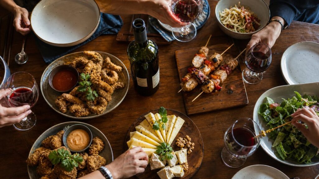 An overhead editorial scene shows a casual spread of fried finger food, grilled skewers, sliced cheese, and an open wine bottle at the center of a crowded table. Warm window light falls evenly across the dishes, with subtle red reflections in the glassware, suggesting a lively, balanced gathering.