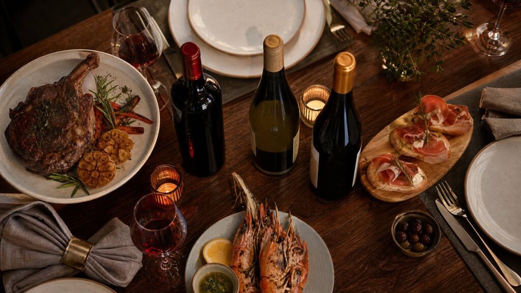 Overhead view of a neatly set dinner table with three different wine bottles arranged near plates of roasted meat, seafood, and light appetizers, suggesting pairing choices before the meal begins. Warm ambient light and subtle crimson reflections in the glass create a refined, practical atmosphere focused on selection.