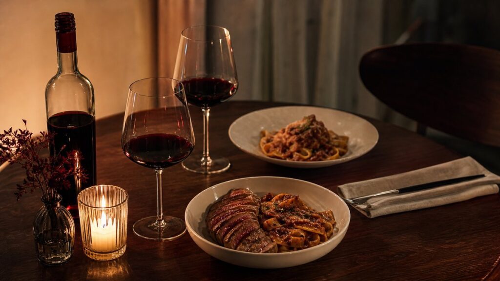 A warmly lit dining table set for two, with a single opened bottle of deep ruby wine beside a candle, linen napkin, and two partially served plates of rich pasta and seared meat. The scene feels intimate and thoughtful, with soft shadows, subtle crimson reflections, and a refined restaurant-at-home mood.