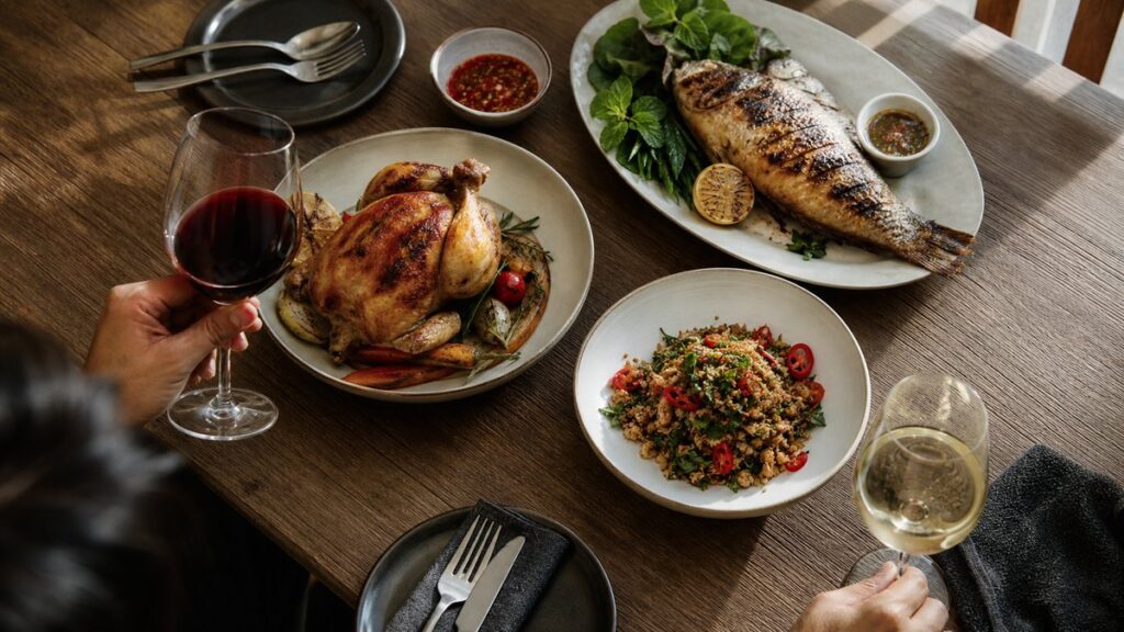 An overhead process scene of a wooden dining table set for pairing wine with food: a roasted chicken dish, grilled fish, a spicy herb plate, and two different wine glasses arranged in practical order. Natural window light falls across the spread, with calm spacing and understated red accents in the meal.