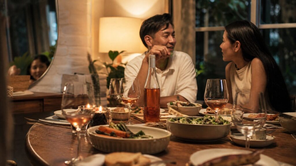 A cozy home dining table is captured in a wide environment shot, where a rosé bottle sits centered among shared dishes, candles, and half-filled glasses after a casual meal. Evening light from a nearby window blends with soft lamp glow, giving the room a memorable, intimate feeling without looking staged.