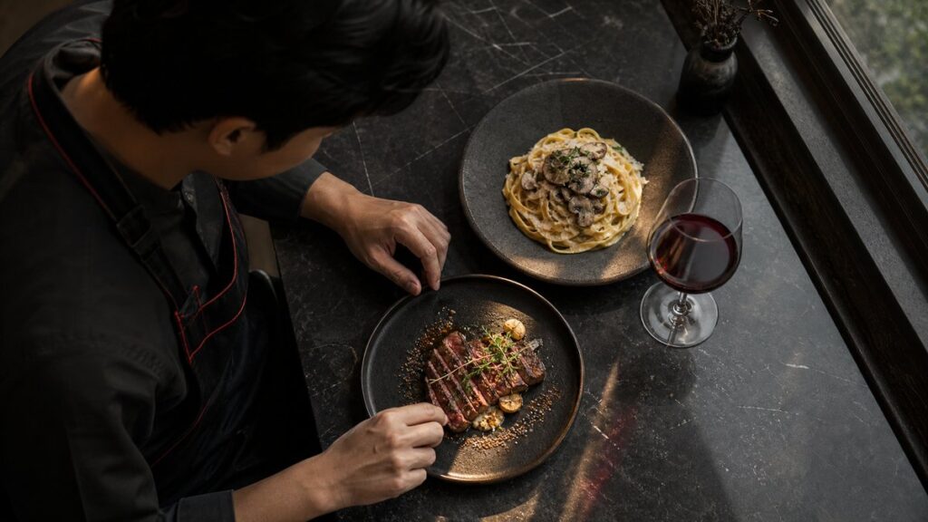 Overhead view of a chef’s table with a plated steak, mushroom pasta, and a red wine glass arranged in careful balance on dark stoneware. Natural window light falls across the food, highlighting texture and color contrast, while the composition feels elegant, deliberate, and quietly luxurious without looking staged.