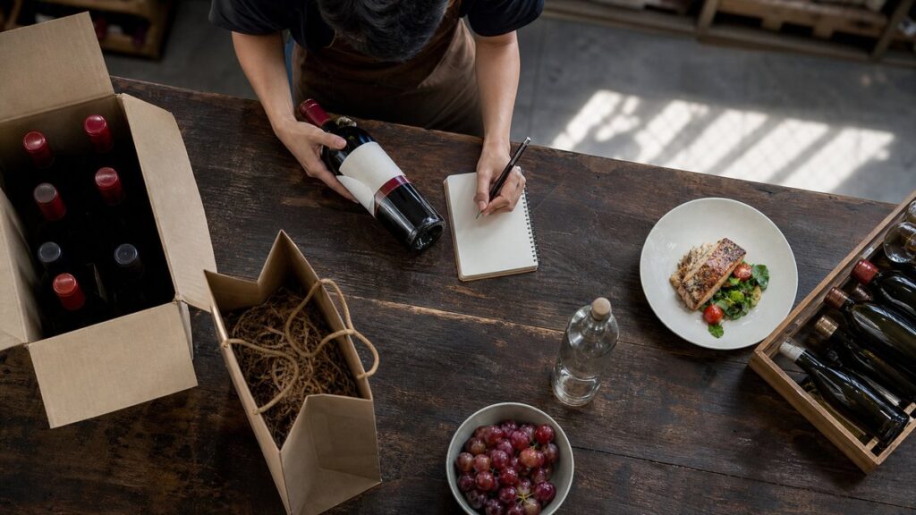 Overhead view of an open wine shop counter where one bottle is being compared with a handwritten budget note, a gift bag, and a simple dinner plate nearby. Natural daylight falls across the arrangement, and the scene feels practical and composed, with muted red accents in the bottle neck and fruit nearby.