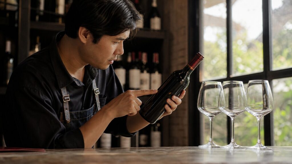A close hand action scene shows one hand gently rotating a wine bottle over a counter while the other points toward the neck and fill level, with a few matched glasses waiting nearby. Warm window light and a restrained palette of deep red liquid, black shadows, and soft neutral surfaces create a practical, confident mood.