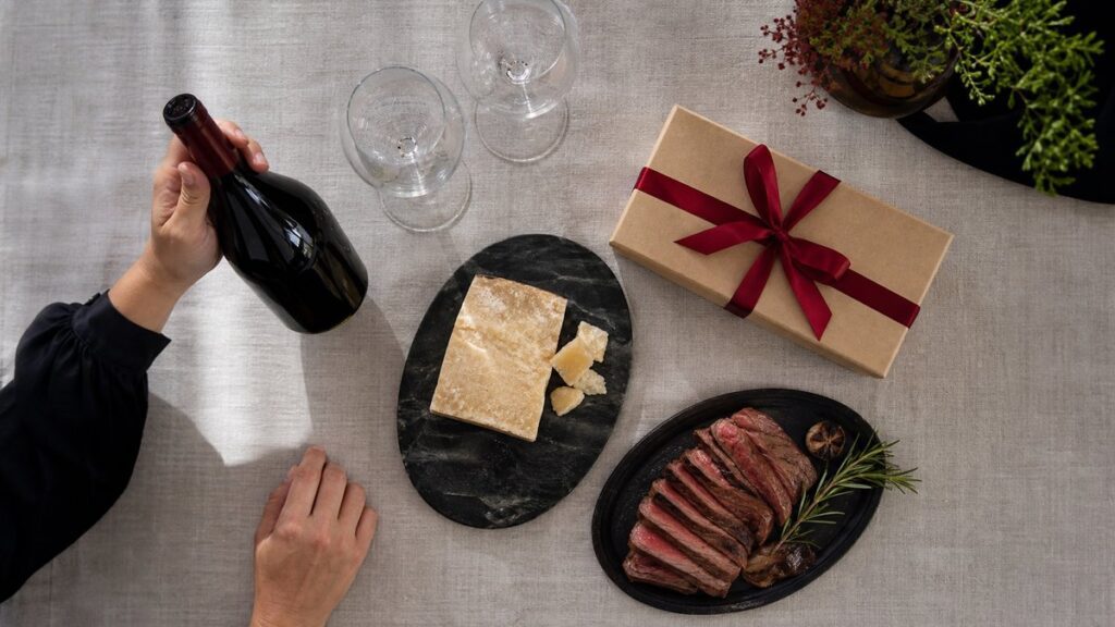 Overhead view of a neatly arranged tasting setup on a linen-covered table: one bottle, two unopened glasses, a piece of aged cheese, sliced steak, and a wrapped gift box tied simply in deep red ribbon. Soft daylight falls evenly across the scene, creating a practical, thoughtful mood of choosing with intention.