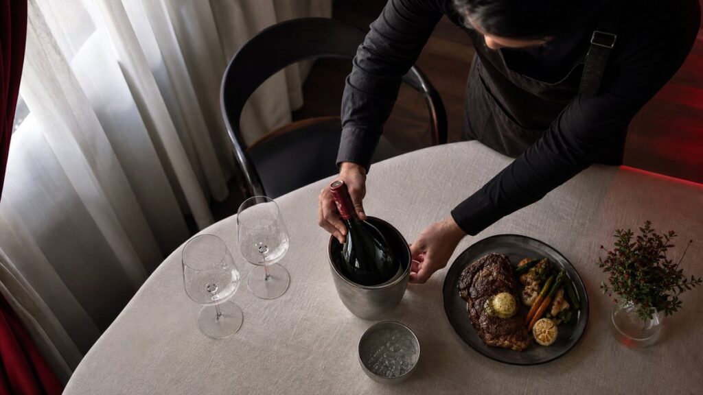 An overhead process scene captures a server arranging a chilled bottle, two proper wine glasses, and a steak dinner on a linen-covered table, with a small bowl of ice water nearby for temperature control. The setting feels refined and quiet, lit by natural window light with subtle crimson highlights.