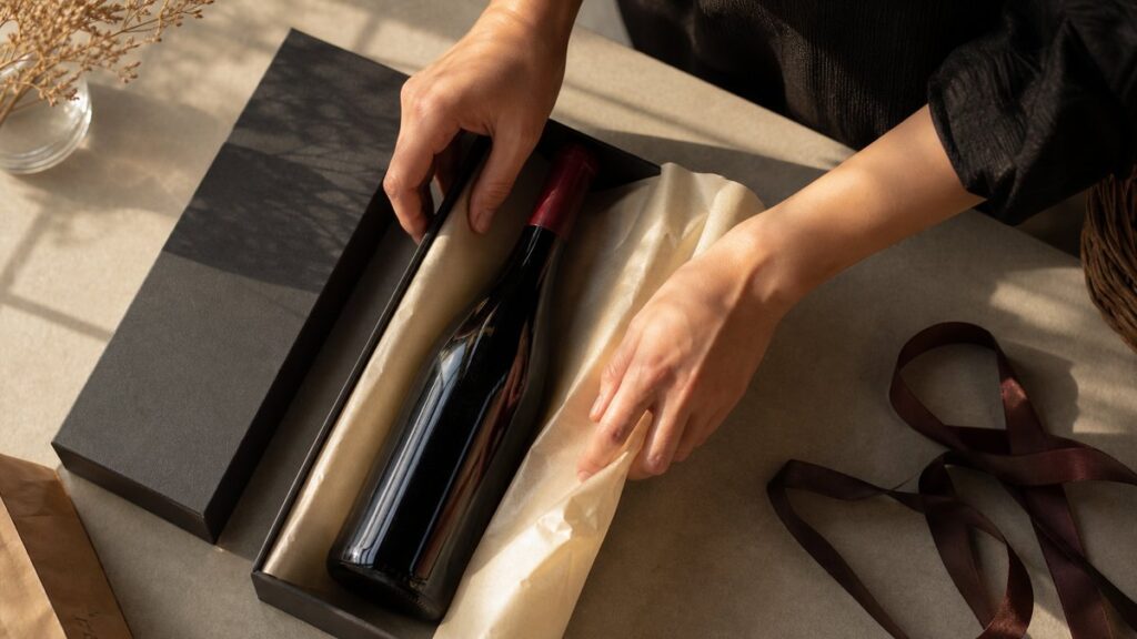 Overhead view of a neatly arranged gift preparation setup: one wine bottle, a matte black box, cream tissue paper, and a dark ribbon laid carefully on a neutral table. Warm side lighting reveals precise hands adjusting the wrapping, giving the scene a polished, intentional, elegant feel.